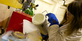 A woman wearing a white coat and protective gloves pours liquid nitrogen from a canister into a funnel.